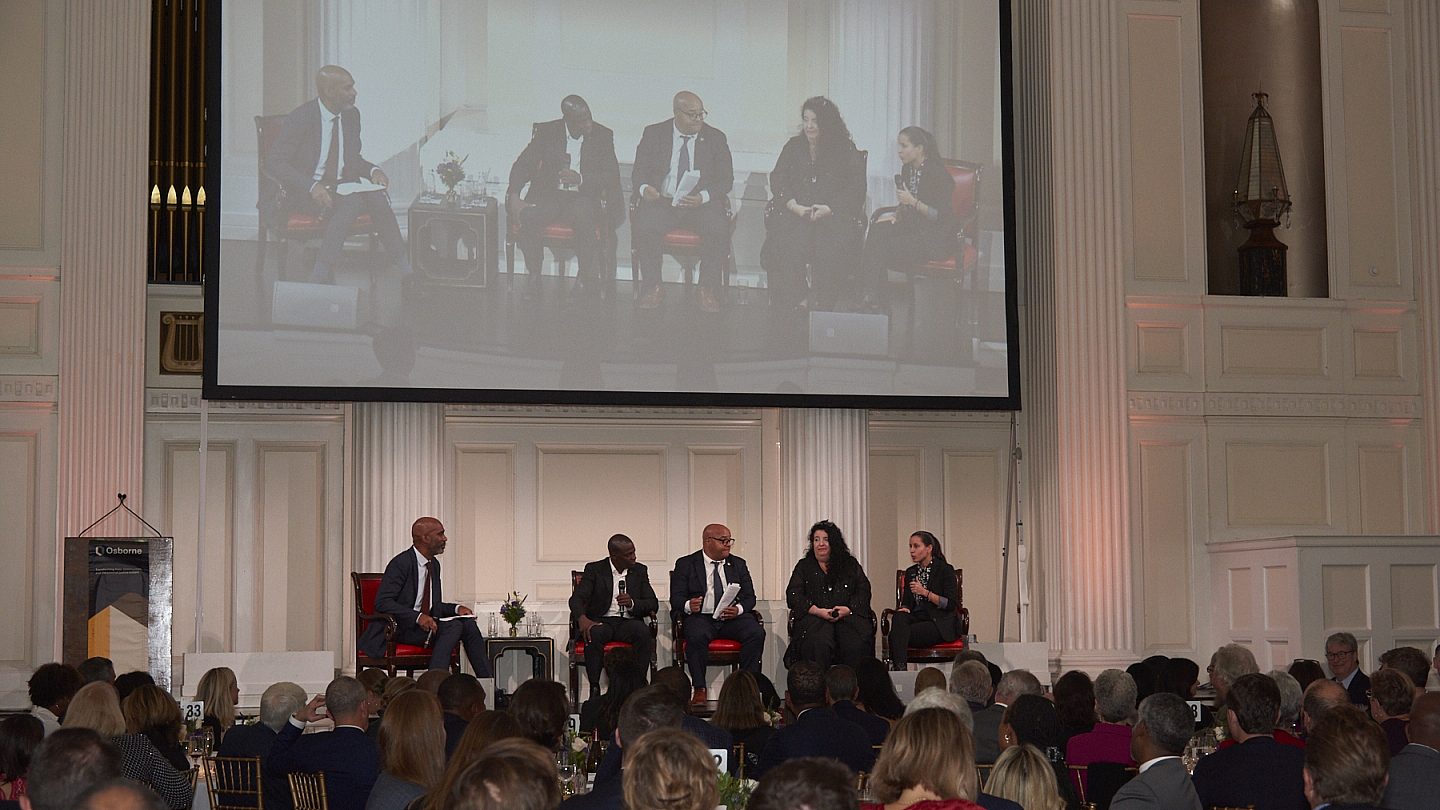 Panel moderator and speakers sitting in a semi-circle together onstage holding microphones. The crowd looks up at them from below.