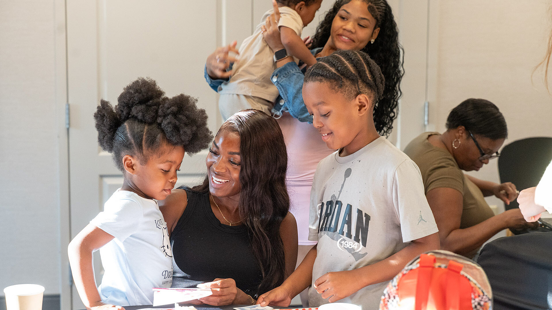 A parent and two kids on either side of them sit at a table making cards. A woman holding a baby is behind them. Another woman sits behind.